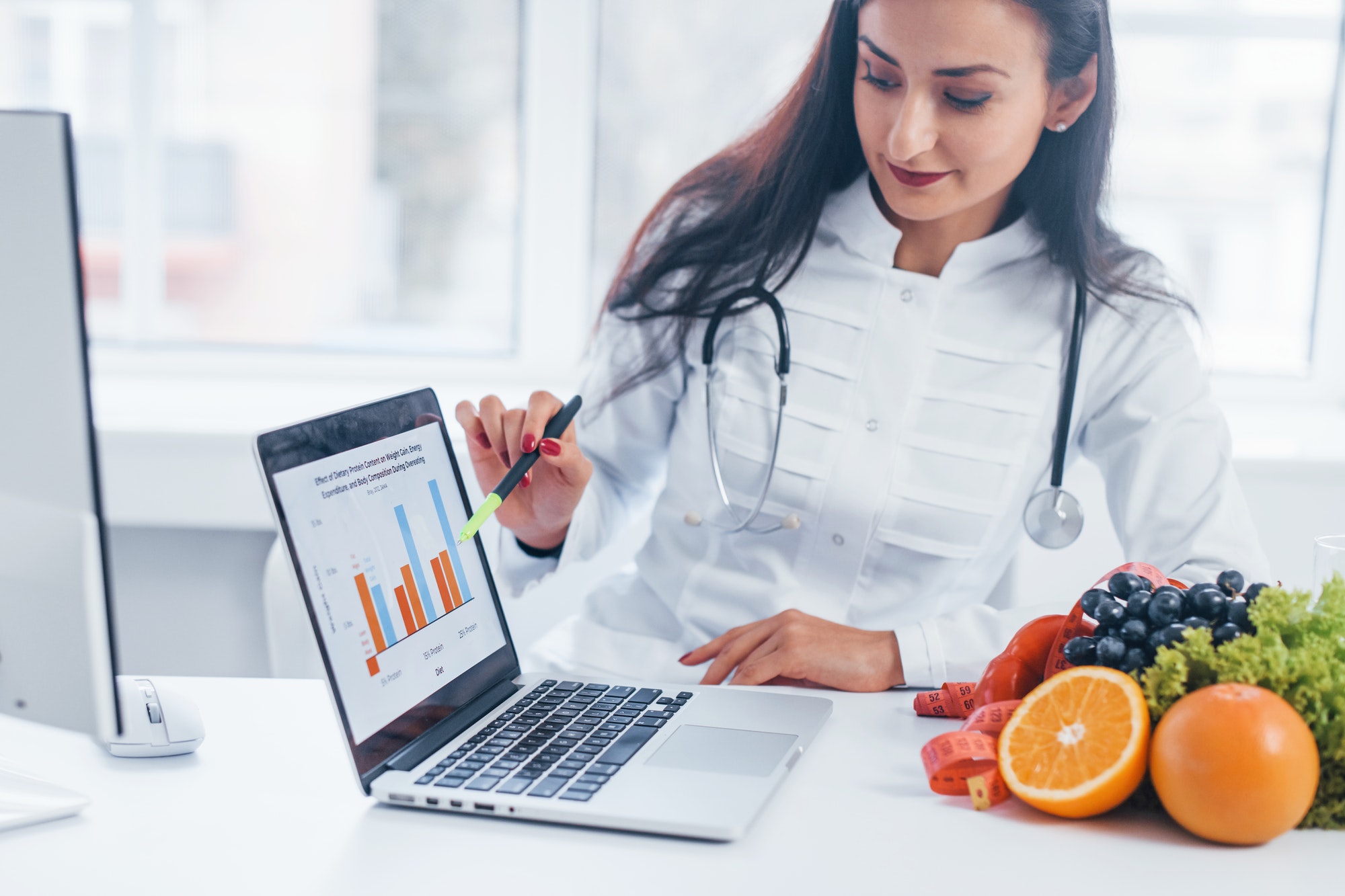 Female nutritionist in white coat sitting indoors in the office at workplace with laptop